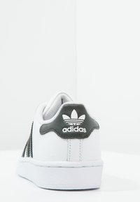 Sneaker bianco con tomaia in pelle testurizzata, linguetta al tallone in pelle verde decorata con il logo Adidas e suola in gomma bianca.