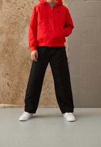 En person i en röd Nike-hoodie, svarta vida byxor och vita sneakers står med händerna i fickorna mot en texturerad vägg.