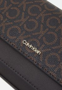Musta rakennepinta oleva lompakko, jossa on ruskea toistuva "CK"-kuvio ja kultainen Calvin Klein -logo, esittelee ompeluyksityiskohtia sileällä nahkasegmentillä.