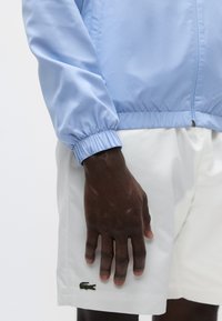 Corta-vento azul claro com punhos elásticos e fecho frontal de correr, combinado com calções brancos com o logotipo do crocodilo verde da Lacoste.