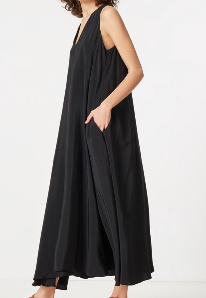 Robe longue noire sans manches avec un profond décolleté en V et des poches latérales portée par une personne debout devant un fond clair uni.