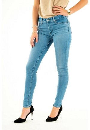 Kvinde iført lyseblå skinny jeans, beige top og sorte højhælede sko med gennemsigtige hæle, står med den ene hånd på hoften og den anden hvilende på låret.