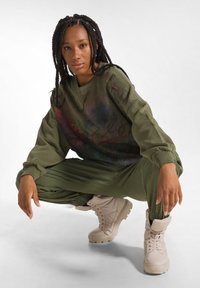 Felpa oversize verde oliva con stampa astratta, abbinata a pantaloni da tuta a gamba affusolata e stivali beige con lacci. Fit e texture rilassati.