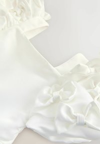 Valge satiinriie, millel on rufflesed varrukad ja kolm kangast paela, luues sujuva ja luksusliku tekstuuri. Suurendatud vaade, mis rõhutab detaile.