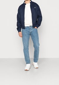 Námořnická bomber bunda s límcem stojáčkem, pod ní bílá mikina, světlé modré džíny a bílé tenisky s béžovými podrážkami. Zelený detail loga na bundě.