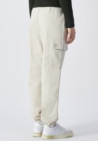 Beige Cargo-Hosen mit einem elastischen Bund und Beinabschlüssen. Sie haben eine Seitentasche und werden mit weißen Sneakern getragen.
