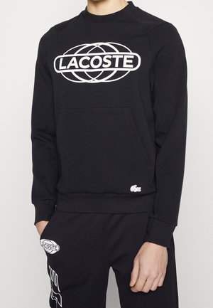 Black sweatshirt featuring a large white Lacoste logo, round neckline, ribbed cuffs, and a front pocket. Additional white croc logo on lower hem.