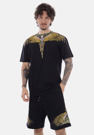 Uomo con capelli ricci e tatuaggi indossa shorts neri coordinati e t-shirt decorati con grandi motivi di ali di uccello gialle e verdi.