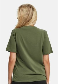 Woman with long blonde hair wearing an olive green short-sleeve t-shirt, shown from the back against a plain white background.