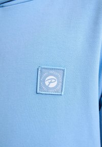 Hanorac albastru deschis cu o textură moale și netedă. Prezintă un patch cu logo pătrată cu text brodat alb, amplasat pe partea din față.