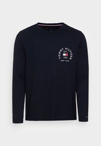 Mekko pitkähihaonen t-paita tummansinisestä pehmeästä puuvillasta, jossa on vasemmassa rinnassa pyöreä valkoinen logo punaisilla ja sinisillä yksityiskohdilla. Pyöreä pääntie.