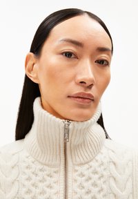 Crèmekleurige kabelgebreide trui met een hoge kraag en een zilveren ritsluiting. De textuur is zacht en chunky, wat het ontwerp accentueert.