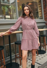 Checked shirt dress in red and white. Features a button-down front, elastic waist, long sleeves, and a tiered skirt. Fabric appears soft.