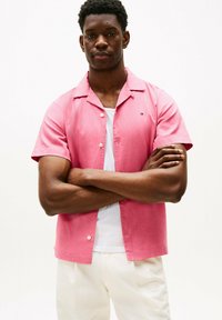 Camicia rosa a maniche corte con colletto a camicia, in tessuto testurizzato, con taschino frontale, indossata sopra una maglietta bianca, abbinata a pantaloni di colore chiaro.