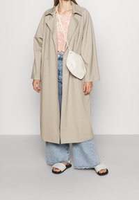 Abrigo largo beige con corte relajado, combinado con una blusa estampada de color rosa claro, jeans de mezclilla anchos azules y sandalias blancas tipo deslizante. Pequeño bolso crema.