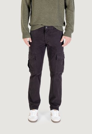 Uomo che indossa pantaloni cargo neri con tasche frontali e laterali, maglione verde oliva e scarpe da ginnastica bianche, in piedi su sfondo grigio chiaro.