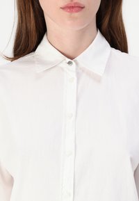 Biela button-up košeľa s klasickým golierom, hladkou textúrou a piatimi prednými gombíkmi, s jednofarebným dizajnom a bez viditeľných vzorov či akcentov.