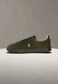 Σκούρο πράσινο sneaker χαμηλού ύψους με λευκό λογότυπο στο πλάι και τη μάρκα "Ralph Lauren" στη σόλα και στη φτέρνα, σε φωτεινή επιφάνεια.