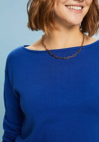 Blauer Strickpullover mit weitem Ausschnitt und gerippter Struktur. Eine goldene Gliederkette betont den Hals. Der Hintergrund ist hellblau.