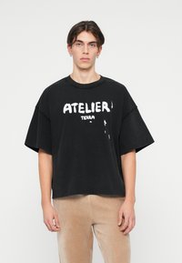 Črna prevelika bombažna majica s kratkimi rokavi, ki ima izrazit bel napis "ATELIER TERRA" in uničen izgled tkanine.