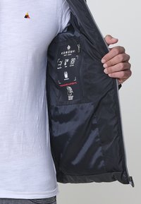 Indvendig i den sorte jakke er der en glat tekstur, plejemærker og et logo. En hvid T-shirt er synlig nedenunder. Jakken har lynlåslukning.