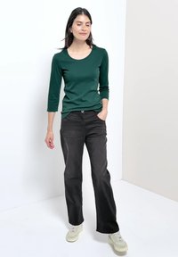 Femme debout portant une chemise verte à manches longues, pantalon noir à jambes larges, baskets blanches, une main dans la poche, sur fond uni.