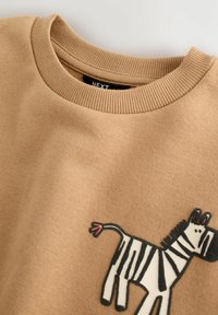 Beige huppari, jossa on ribbetty pyöreä kaula-aukko ja sarjakuvamainen seepra -graafinen kuva edessä olkapään lähellä.