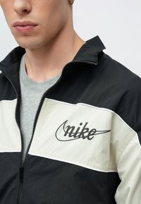 Μαύρο και κρεμ μπουφάν με φερμουάρ και λογότυπο Nike στο στήθος. Διαθέτει ψηλό γιακά και ραφές σε αντίθεση. Φορεμένο πάνω από γκρι μπλούζα.
