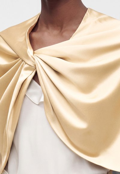 Personne à la peau foncée portant un châle en satin beige lisse et brillant drapé sur un vêtement blanc, avec un nœud torsadé près du cou.