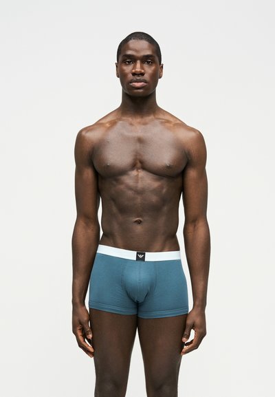 Μυώδης άνδρας που στέκεται μπροστά φοράει βερμούδα boxer teal με λευκή μέση, σε απλό ανοιχτό φόντο.