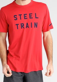 Hombre que lleva una camiseta roja de manga corta con "STEEL TRAIN" en letras negras, combinada con pantalones cortos de color gris oscuro.