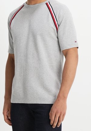 Mężczyzna ubrany w jasnoszarą krótką koszulkę z czerwonymi, białymi i czarnymi paskami na szwach ramion oraz ciemne spodnie.