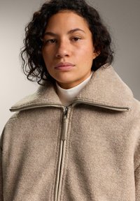 Bež flis jakna z visokim ovratnikom, izpostavljeno zadrgo in teksturirano površino. Mehka tkanina z udobnim krojem in minimalističnim dizajnom.