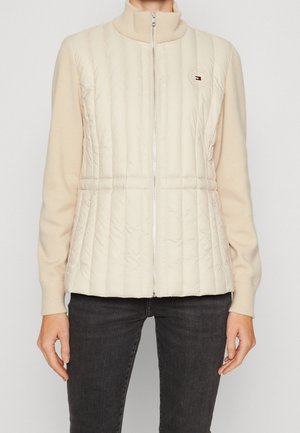 Person iført beige quiltet lynlåsjakke med ribbekant på krave og ærmer, kombineret med mørkegrå jeans, stående mod en lys baggrund.