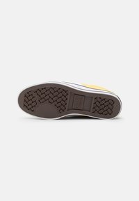 Semelle de chaussure avec une base en caoutchouc brun, dotée d'un motif de semelle texturé, d'un bord blanc et d'un accent jaune près du talon.