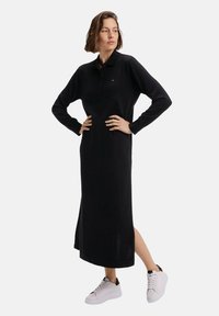 Robe polo noire à manches longues avec un col, texture côtelée, fentes latérales et coupe décontractée, associée à des baskets blanches avec des accents noirs.