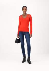 Röd långärmad topp med utskärning på axlarna, kombinerad med marinblå smala jeans. Modellen bär svarta ankelstövlar och en liten svart handväska.