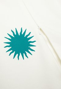 Tissu blanc avec un motif de soleil turquoise aux rayons pointus ; comprend un détail de fermeture éclair. Texture lisse, design simple.