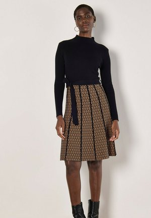 Maglia nera a costine con maniche lunghe, collo alto e dettaglio con fiocco; gonna a trapezio con un motivo geometrico in oro e nero. Stivali neri alla caviglia.