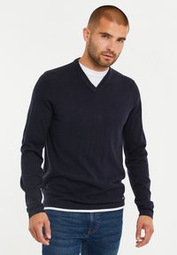 Granatowy sweter z dekoltem w serek, wykonany z gładkiego, dzianinowego materiału z długimi rękawami, noszony na białej koszuli. Posiada ściągacze przy mankietach i u dołu.