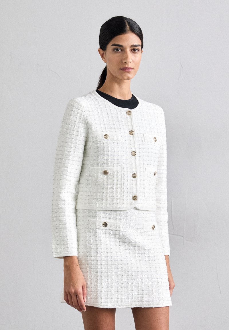 Veste en tweed blanc avec boutons dorés, motif texturé, coupe courte et manches longues. Associée à une mini-jupe assortie.