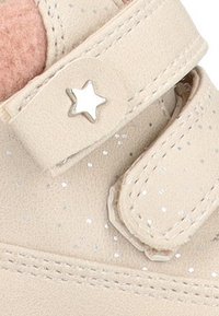 Zapato de cuero beige con una correa de velcro, que presenta un acento de estrella plateada y pequeños detalles brillantes en el material. Forro de tela rosa suave.