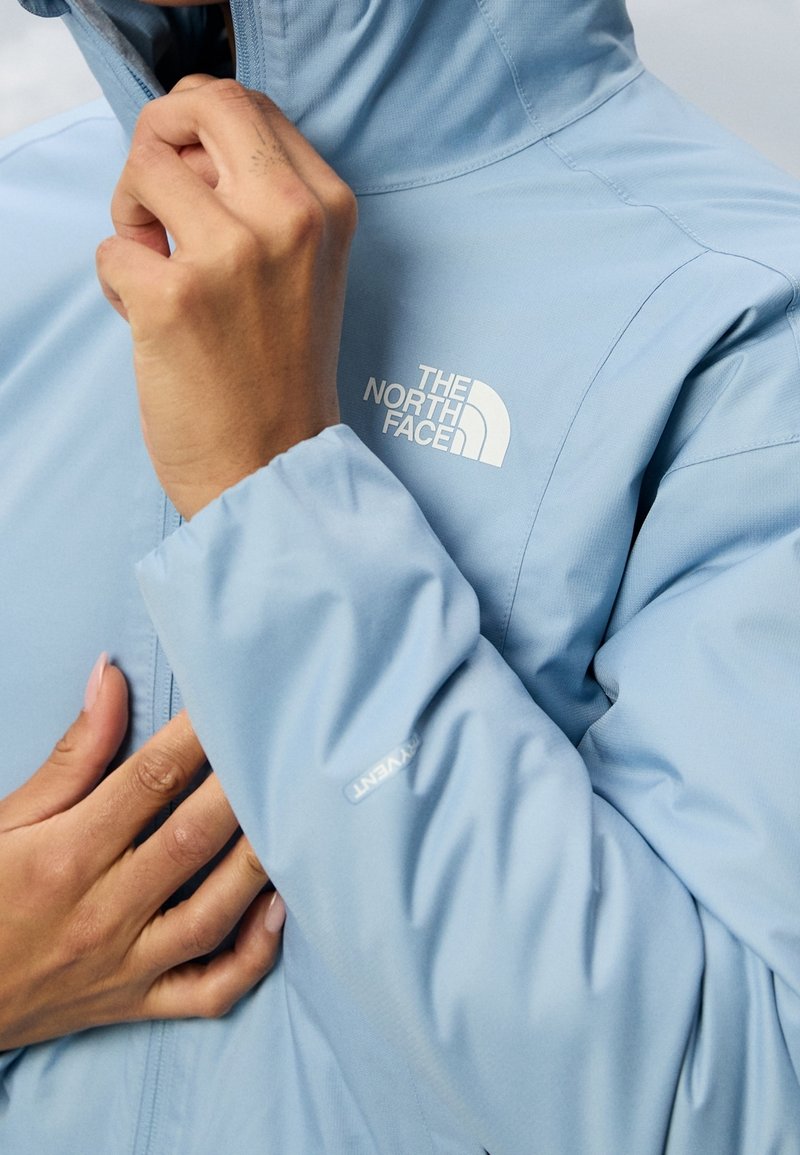 Jasnoniebieska wodoodporna kurtka z wysokim kołnierzem, zamkiem błyskawicznym i wyraźnym białym logo. Gładka tekstura i elastyczne mankiety.