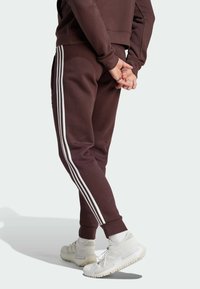 I pantaloni della tuta marroni presentano strisce laterali bianche, caviglie regolabili e una vestibilità ampia. Indossati con scarpe da ginnastica bianche e calze texturizzate.