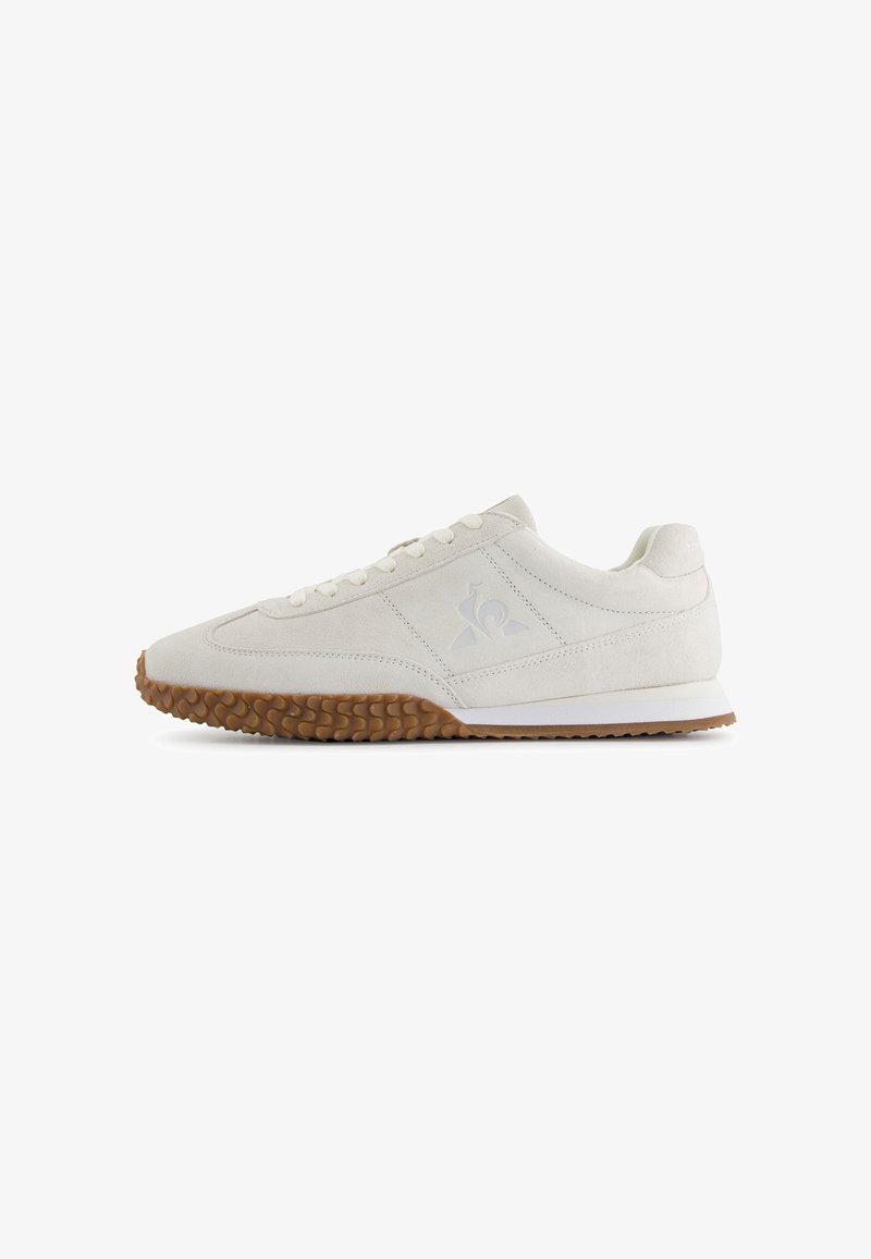 Baskets en suede beige à profil bas avec semelle en gomme, lacets blancs et logo subtil sur le côté, montrées de côté sur fond blanc.