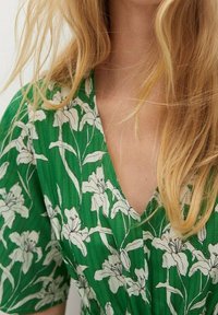 Grünes Kleid aus strukturiertem Stoff mit einem floralen Muster in Weiß. Das Design beinhaltet einen V-Ausschnitt und kurze Ärmel.