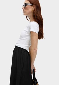 Witte fitted t-shirt gecombineerd met een zwarte, geplooide rok. Accessoires zijn rechthoekige zonnebrillen en een zwarte, gestructureerde handtas.