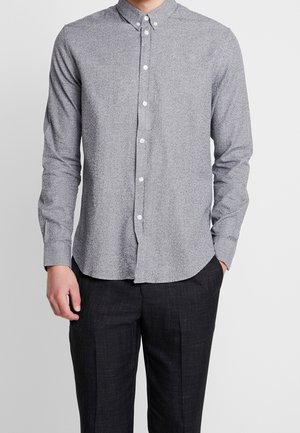 Mann trägt ein graues, langärmliges Hemd mit Knöpfen und einem Kragen mit Knopfverschluss sowie schwarze Hose, eine Hand in der Tasche.