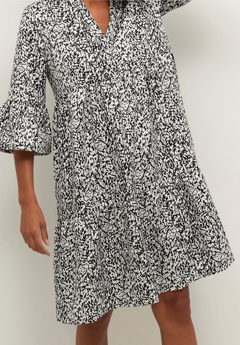 Robe à motifs noir et blanc avec un col en V, coupe ample et manches cloche à trois quarts. Elle présente une texture froncée et un ourlet à volants.