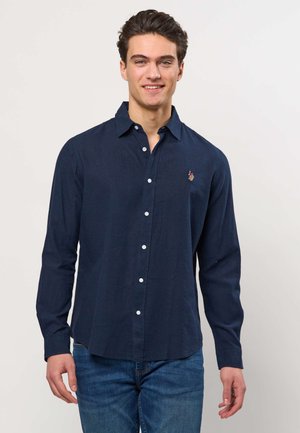 Noormees naeratab, kannab mereväevärvi sinist nööbitavat särk ja siniseid teksapükse, seisab ühevärvilise tausta ees.